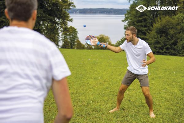 Junge Männer spielen Tischtennis im Freien auf einer Wiese mit Blick auf einen See.