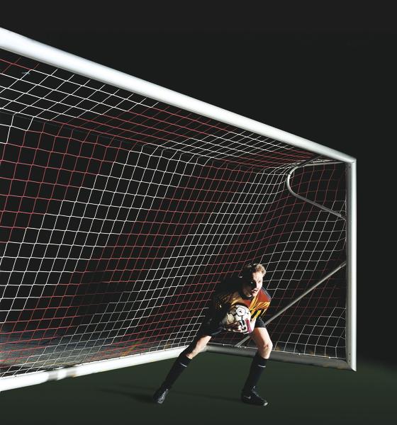 Torwart fängt Fußball vor dem Tor in einem dunklen Stadion mit rotem Netz.