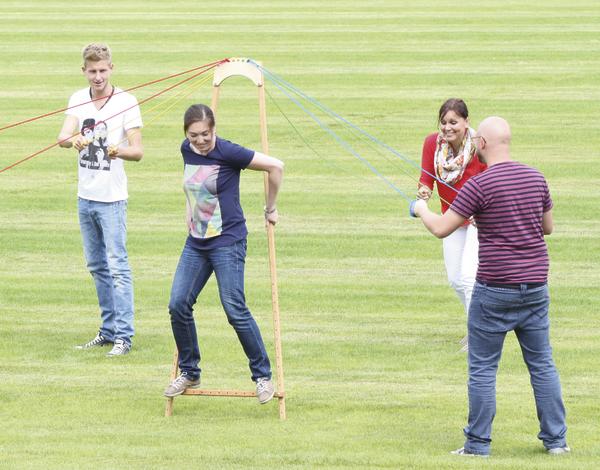 Vier Personen spielen im Freien ein Teamspiel mit Stelzen und Seilen auf einer grünen Wiese.