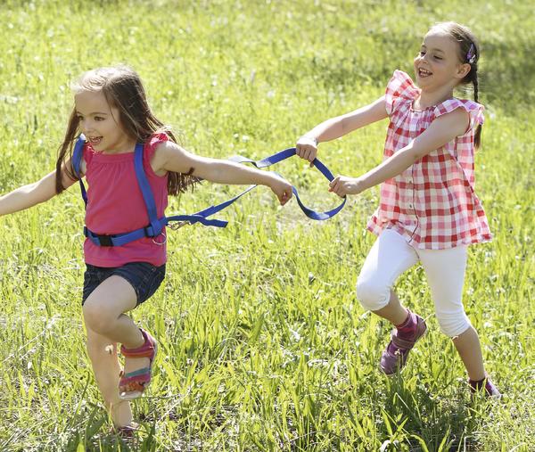 Zwei Mädchen spielen lachend mit Sicherheitsgeschirr auf einer grünen Wiese im Sommer.