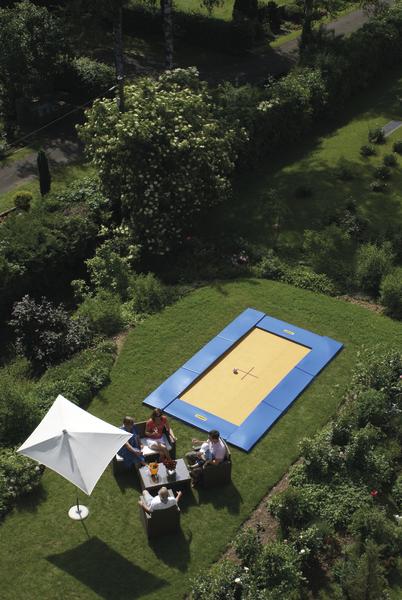 Garten mit rechteckigem Trampolin, Sitzgruppe und Sonnenschirm, umgeben von grünen Pflanzen.