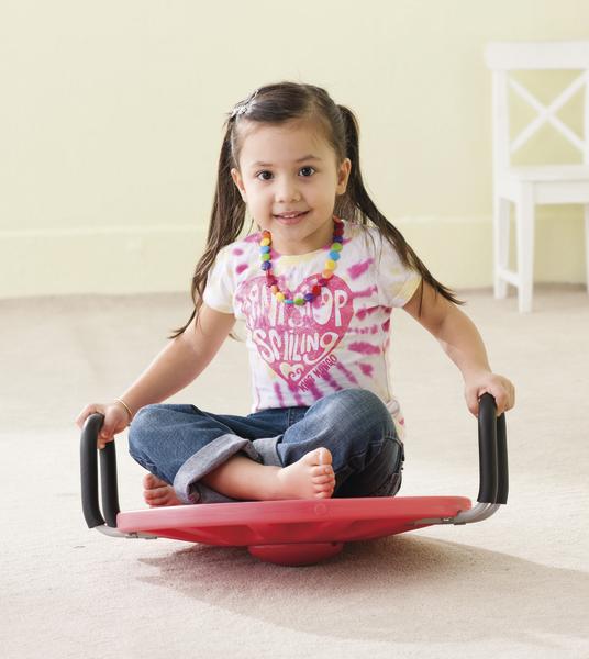 Mädchen sitzt lachend auf rotem Wippbrett im Wohnzimmer, trägt buntes T-Shirt und Jeans.