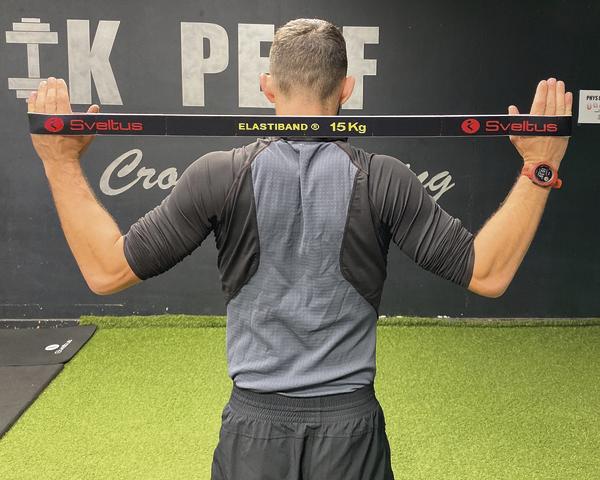 Mann trainiert mit 15 kg Elastikband im Fitnessstudio vor schwarzer Wand mit Schriftzug.