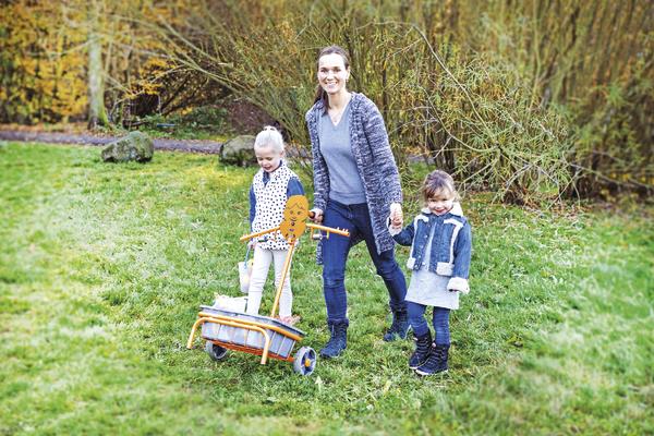 Frau mit zwei Kindern im Park, zieht Wagen mit Holzfigur, herbstliche Natur im Hintergrund.