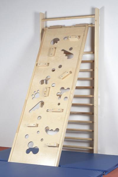 Kletterwand aus Holz mit verschiedenen Griffen und Löchern vor einer Sprossenwand und blauer Matte.