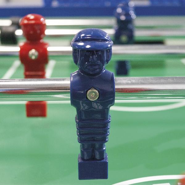 Blaue Kickerfigur auf einer Stange montiert, im Hintergrund weitere Spielfiguren auf dem Spielfeld.