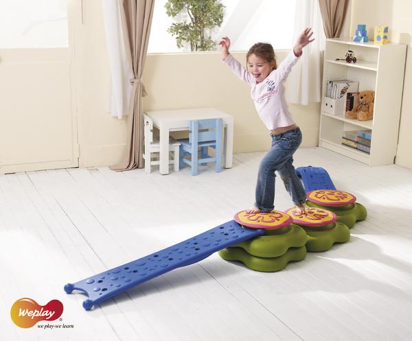Kind balanciert auf buntem Indoor-Spielgerät in hellem Kinderzimmer mit Möbeln und Spielzeug.
