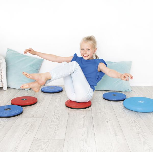 Mädchen balanciert auf bunten Balancekissen im hellen Raum mit Kissen im Hintergrund.