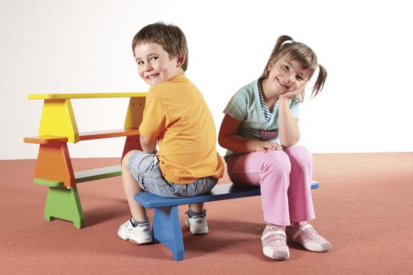 Junge und Mädchen sitzen lachend auf bunter Kinderbank vor weißem Hintergrund.