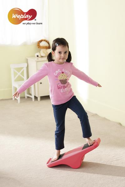 Mädchen balanciert auf rotem Wippbrett im hellen Zimmer mit Spielzeug im Hintergrund.