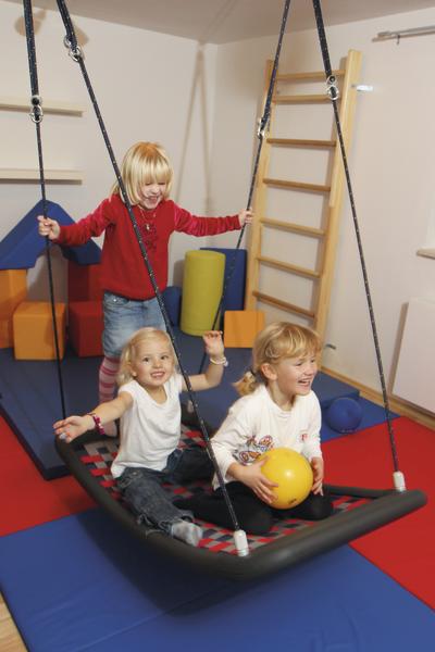 Drei Kinder spielen fröhlich auf einer Hängeschaukel in einem bunten Indoor-Spielraum.