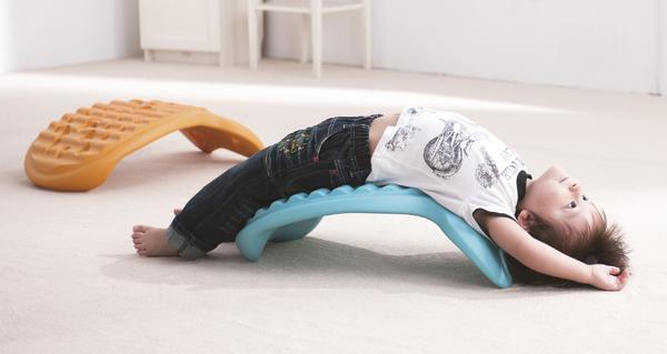 Kind entspannt auf blauem ergonomischem Rückenstützkissen im hellen Raum, Spielgerät im Hintergrund.