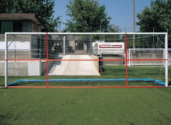Fußballtor mit rotem Trainingsnetz auf grünem Kunstrasen vor Gebäuden und Bäumen.