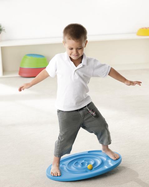 Junge balanciert barfuß auf blauem Wackelbrett mit gelbem Ball in hellem Raum.