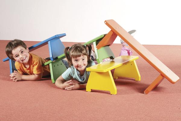 Zwei Kinder liegen lachend auf dem Boden, umgeben von bunten Holzstühlen und einer Wippe.