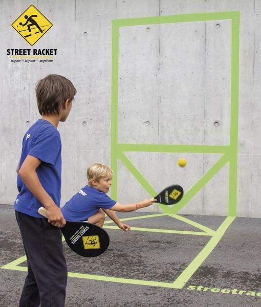 Zwei Jungen spielen Street Racket an einer markierten Wand mit Schlägern und gelbem Ball.