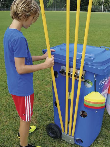 Junge in Sportkleidung nimmt Trainingsstäbe aus blauem Sportwagen auf Fußballfeld.
