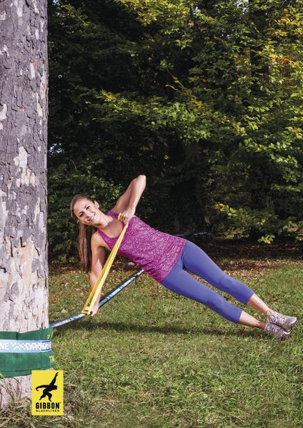 Frau trainiert Balance mit Slackline zwischen Baum und Boden im Park bei Sonnenschein.
