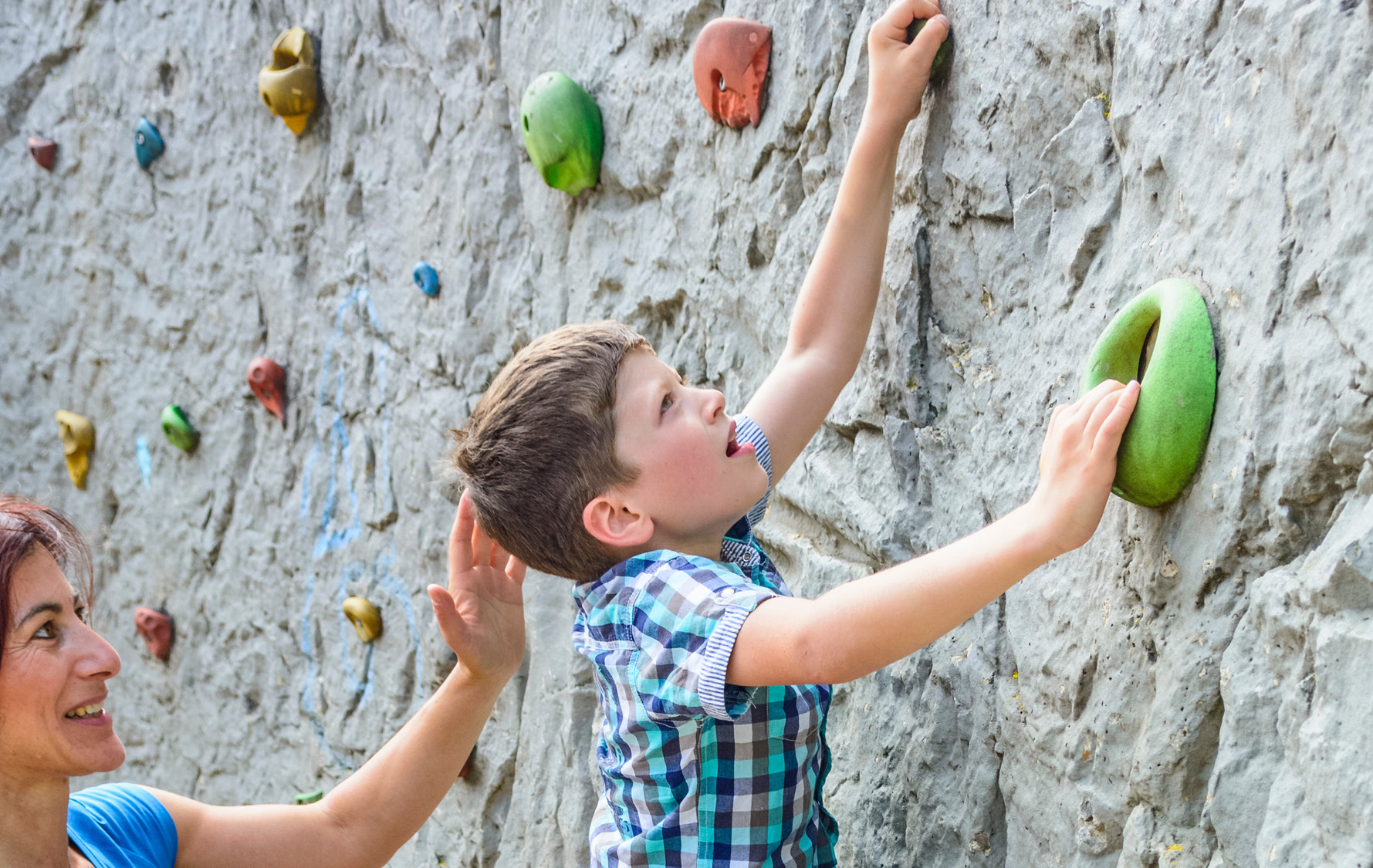 Kinder klettert mit Hilfestellung einer Frau an einer künstlichen Boulderwand im Außenbereich.